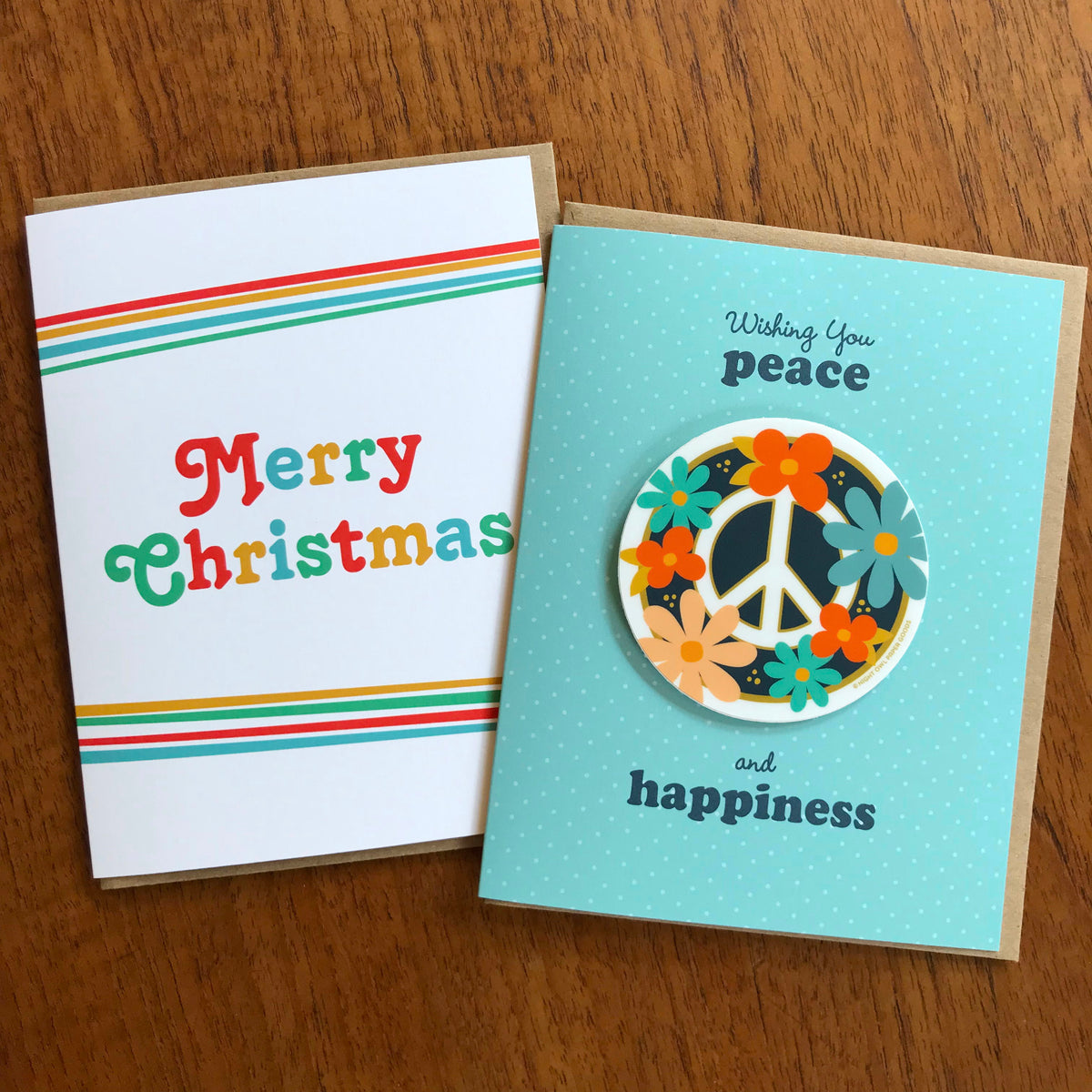 Big Christmas Holiday Card and Floral Peace Sticker Holiday Card shown side by side on a wood surface.