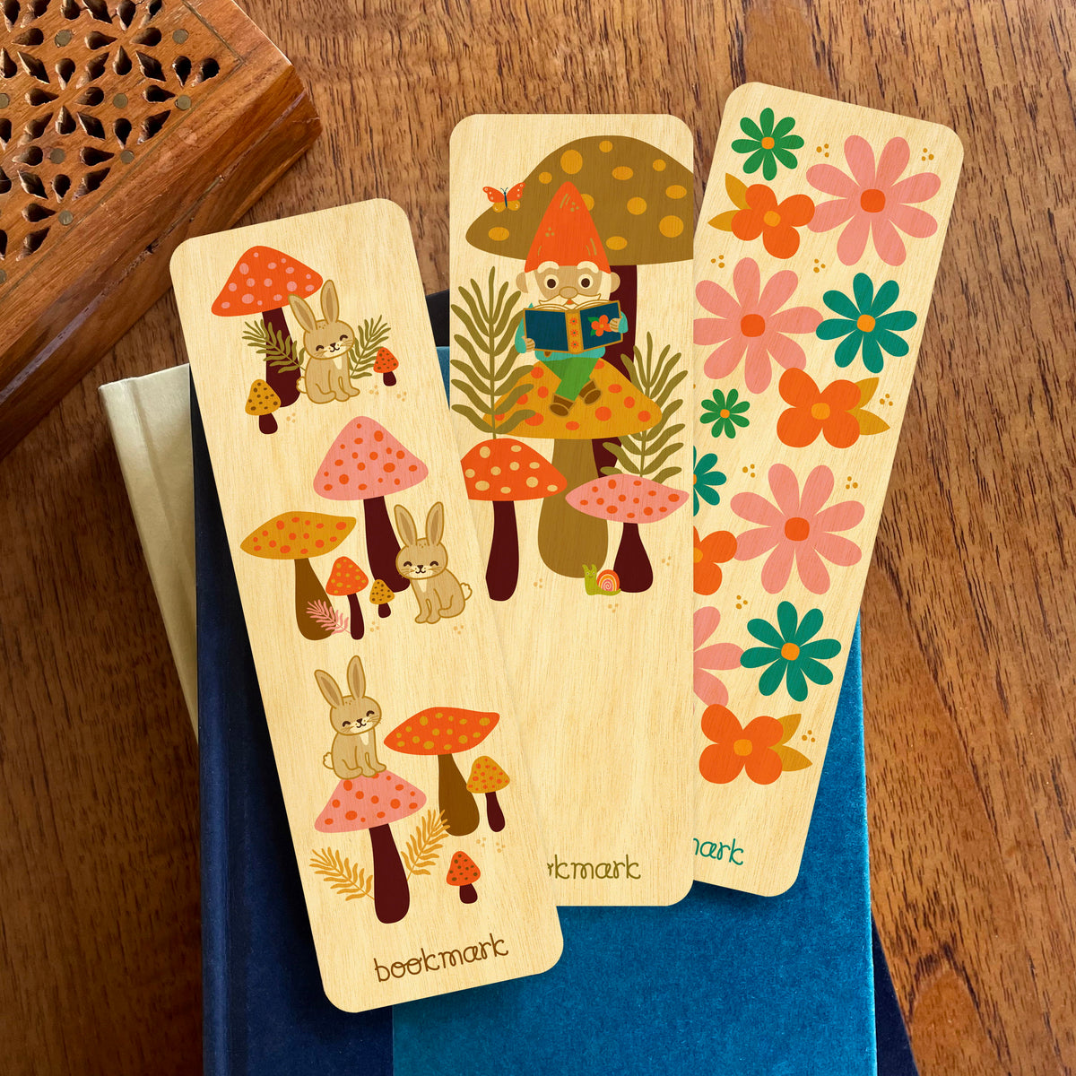 Group photo of three wooden bookmarks resting on a stack of books. Bookmarks feature rabbits, a gnome reading on a mushroom and a colorful flower pattern