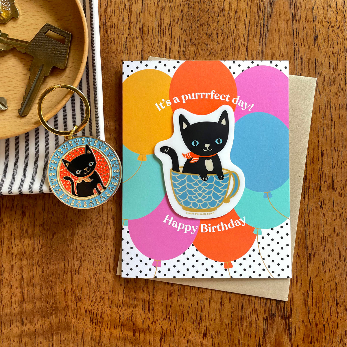 Black cat enamel keychain next to Purrrfect Day Sticker Birthday Card