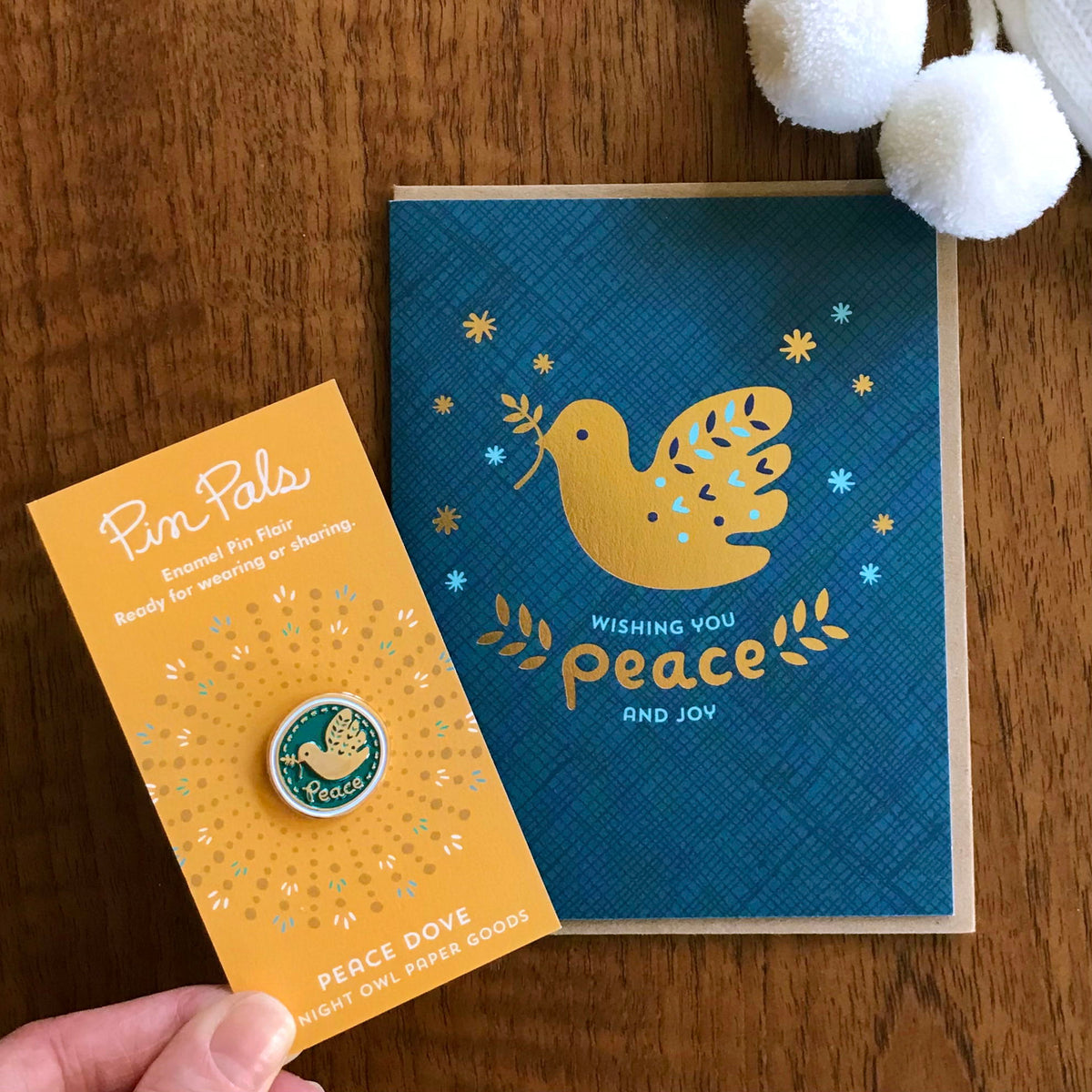 Blue and gold dove enamel pin on packaging text reads 'peace' and a blue holiday greeting card with a gold foil dove and text 'Wishing you peace and joy' 