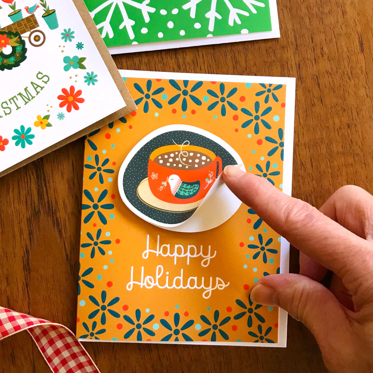 Holiday Cocoa Sticker Holiday Card featuring a removable vinyl sticker of a red mug of hot cocoa with marshmallows and a teal bird design on a warm gold background with teal flowers and the greeting "Happy Holidays." Shown with a white envelope. A finger lifts the edge of the sticker to demonstrate that it's removable.