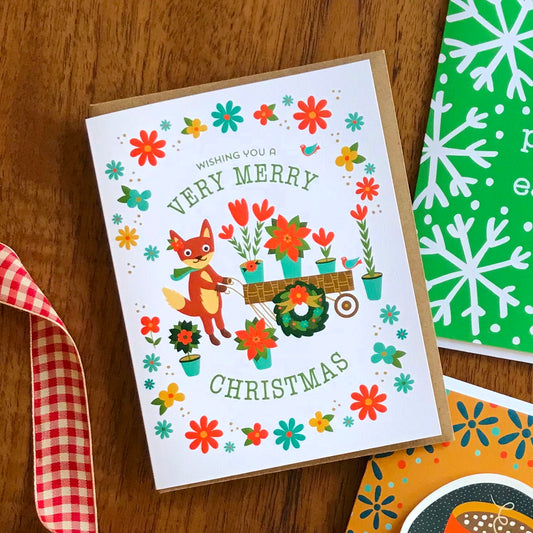 Foxy Christmas folded paper holiday card featuring a fox pulling a cart of poinsettias and potted flowers surrounded by a wreath of colorful blooms and birds with the greeting "Wishing You a Very Merry Christmas." Shown with a kraft envelope.
