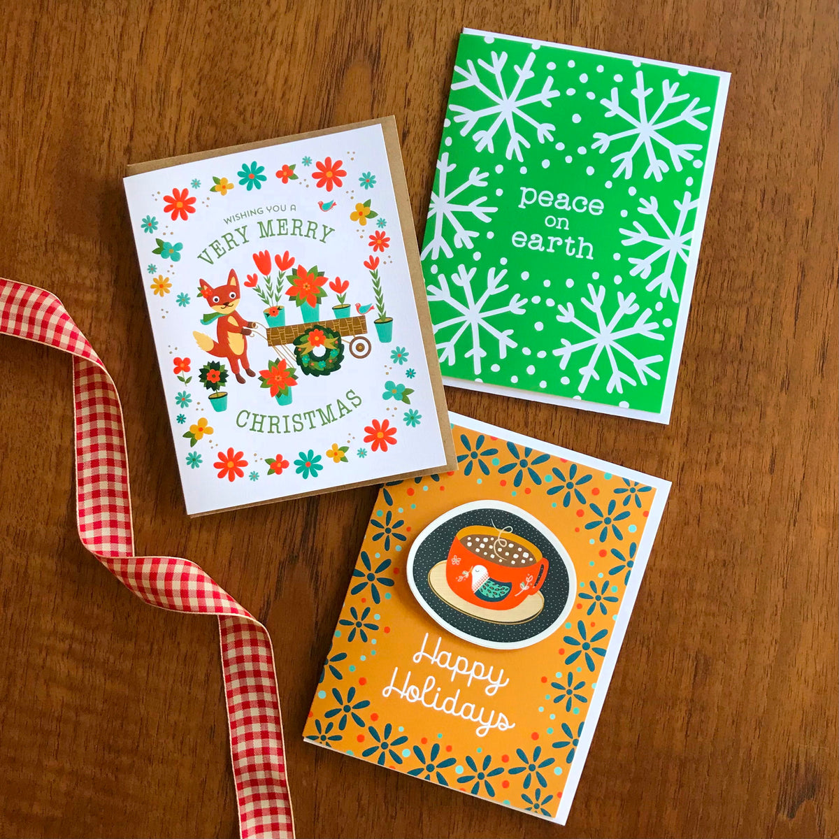 Foxy Christmas, Green Peace and Cozy Christmas holiday cards shown laying on a wood tabletop.