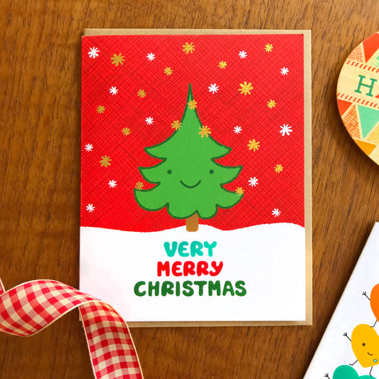Christmas card with a smiling green tree with red background and gold foil snowflakes. Text reads 'Very Merry Christmas'
