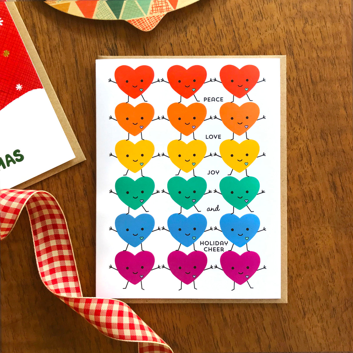 United Hearts folded paper holiday card featuring rows of smiling rainbow hearts with arms and legs holding hands in red, orange, yellow, green, blue and purple with the greeting "Peace Love Joy and Holiday Cheer." Shown with a kraft envelope on a wood tabletop.