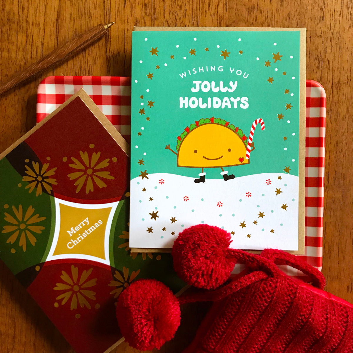 Taco Gold Foil Holiday Card featuring a smiling taco in black boots holding a candy cane against a snowy aqua background with gold foil stars and a "Wishing You Jolly Holidays" greeting. Shown with a kraft envelope next alongside another holiday card, atop a a festive checkered red background.