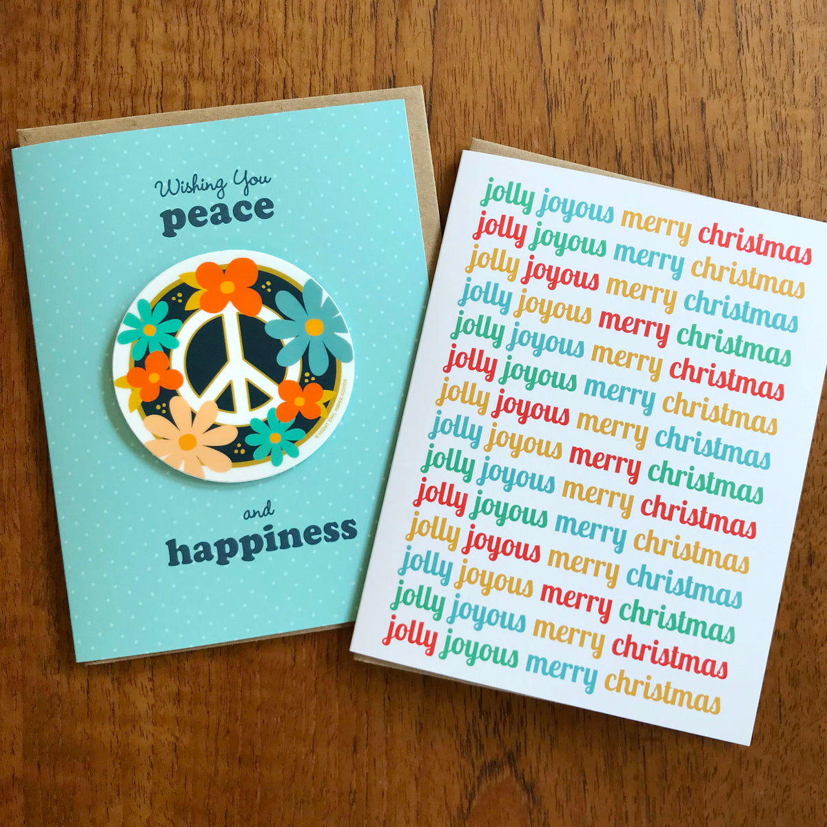 Jolly Joyous and Floral Peace folded holiday cards side by side on a wood table.