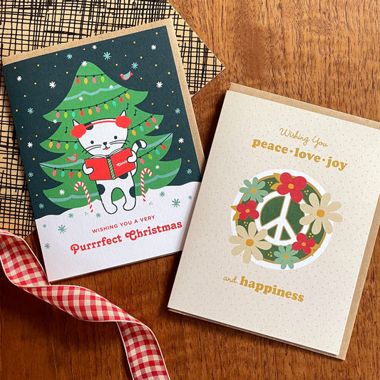 Caroling Kitty and Blooming Peace Cream folded paper holiday cards pictured aside one another on a wood table with holiday ribbon decor. Both are paired with kraft envelopes.