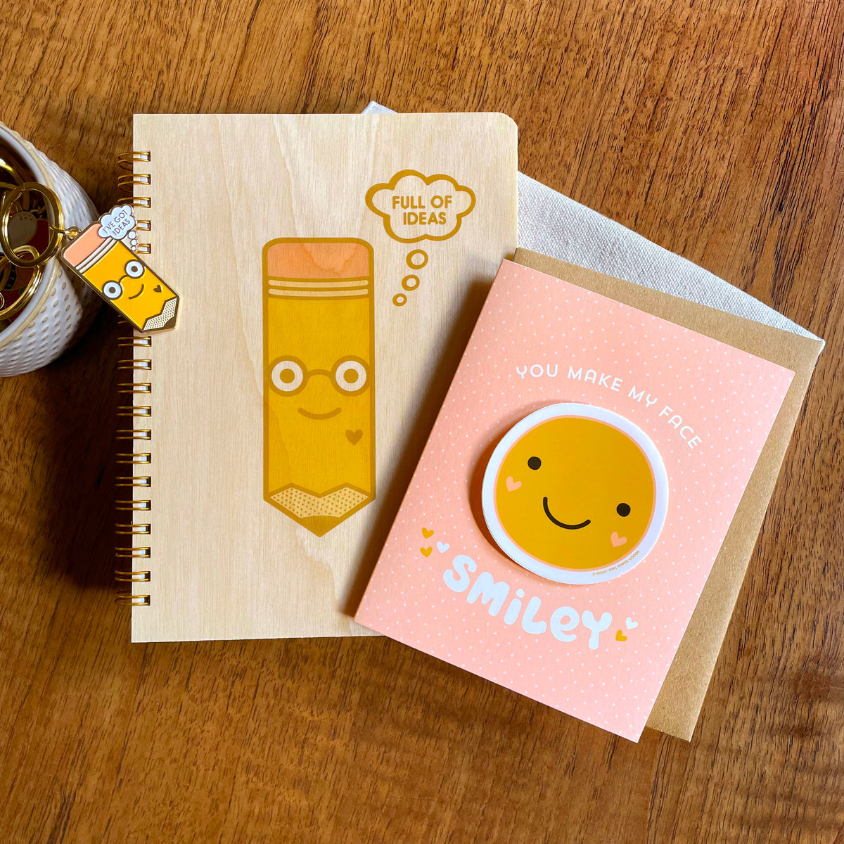 Full of Ideas Wood Journal, Idea Pencil Enamel Keychain and Smiley Face Sticker Card side-by-side on a wood table.