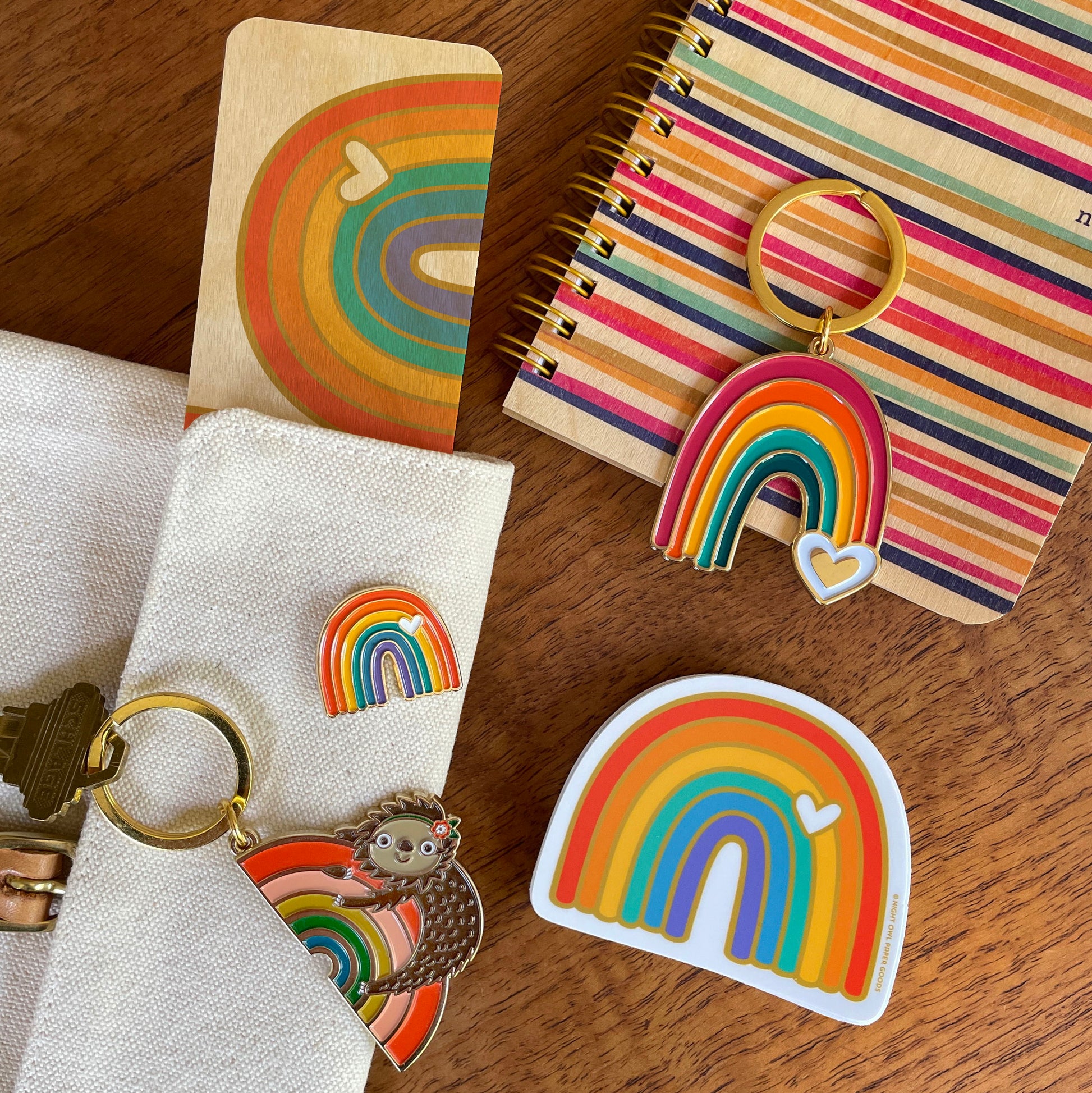 Rainbow enamel keychain displayed alongside an assortment of rainbow-themed products, including a Rainbow enamel pin, Rainbow Sloth keychain, Rainbow bookmark, Rainbow sticker and Party Lines notebook.