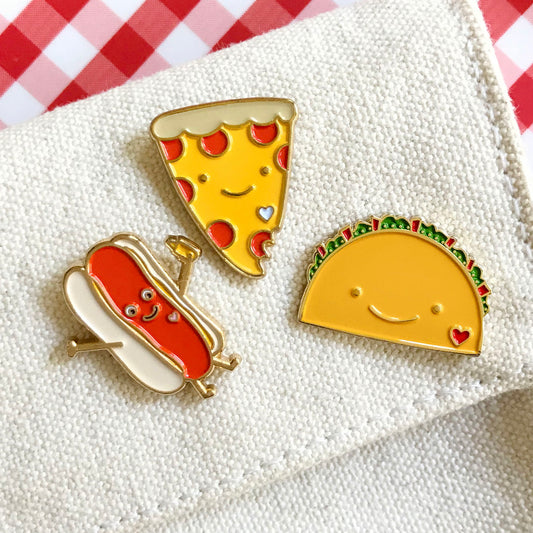 Three food lover enamel pins hot dog pizza and taco. Each pin is personified with a simple smile and small heart shape detail.