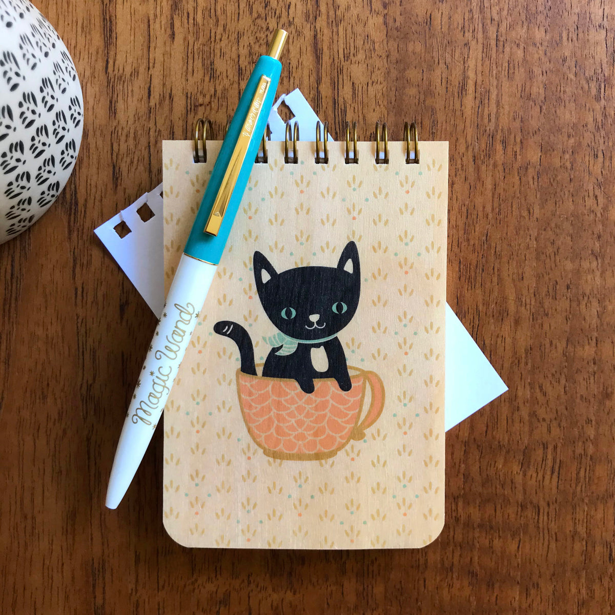 Spiral-bound wood mini notepad with birch wood covers featuring a black cat with green eyes sitting inside a pink teacup alongside an aqua and white Magic Wand ballpoint pen.
