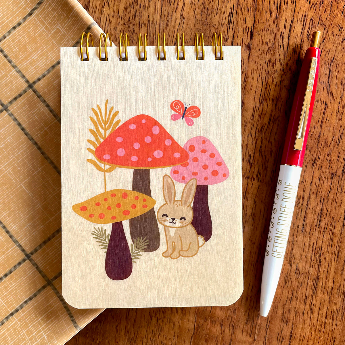 Spiral-bound wood mini notepad with birch wood covers featuring a happy bunny nestled among spotted mushrooms with a butterfly and wildflowers alongside a Getting Stuff Done ballpoint pen.