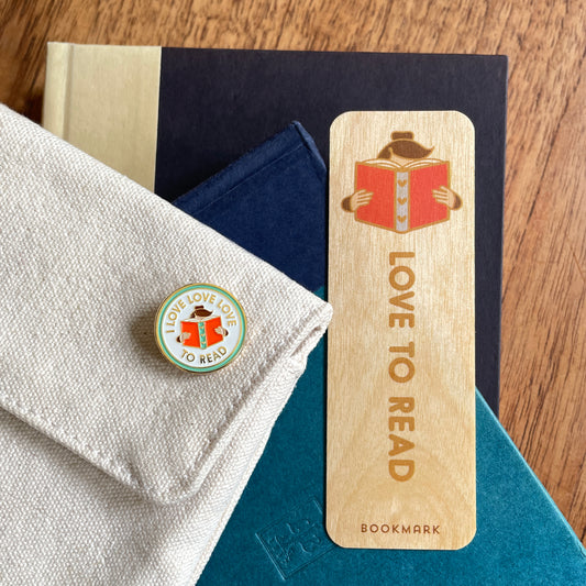 A wooden bookmark featuring a woman reading a book and the words 'LOVE TO READ' displayed on top of several books, alongside a matching round enamel pin.