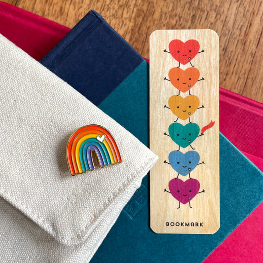 A wooden bookmark with six colorful smiling hearts and the word 'BOOKMARK' at the bottom, displayed next to a rainbow enamel pin on a folded white fabric surface with books in the background.