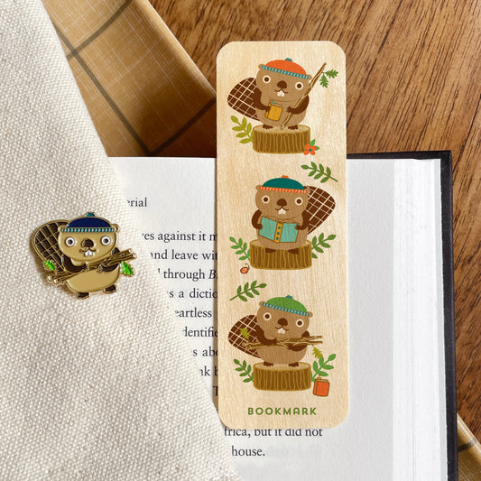 A wooden bookmark featuring three beavers each holding a book, displayed on an open book next to a matching beaver enamel pin on a textured cream fabric.