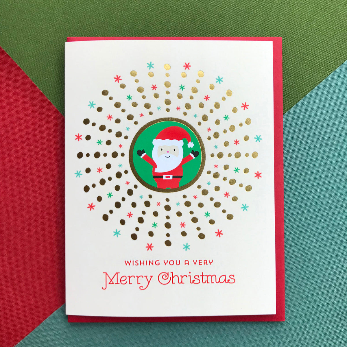 Santa Gold Foil Holiday Card featuring a cheerful Santa Claus in a green circle surrounded by a gold foil dot sunburst and red and green snowflakes on a cream background with the greeting "Wishing You a Very Merry Christmas." Shown with a red envelope.
