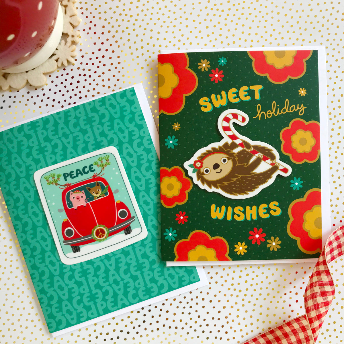 Peace Pals Sticker Holiday Card and Sweet Sloth Sticker Holiday Card sitting side by side on a festive gold dotted surface.
