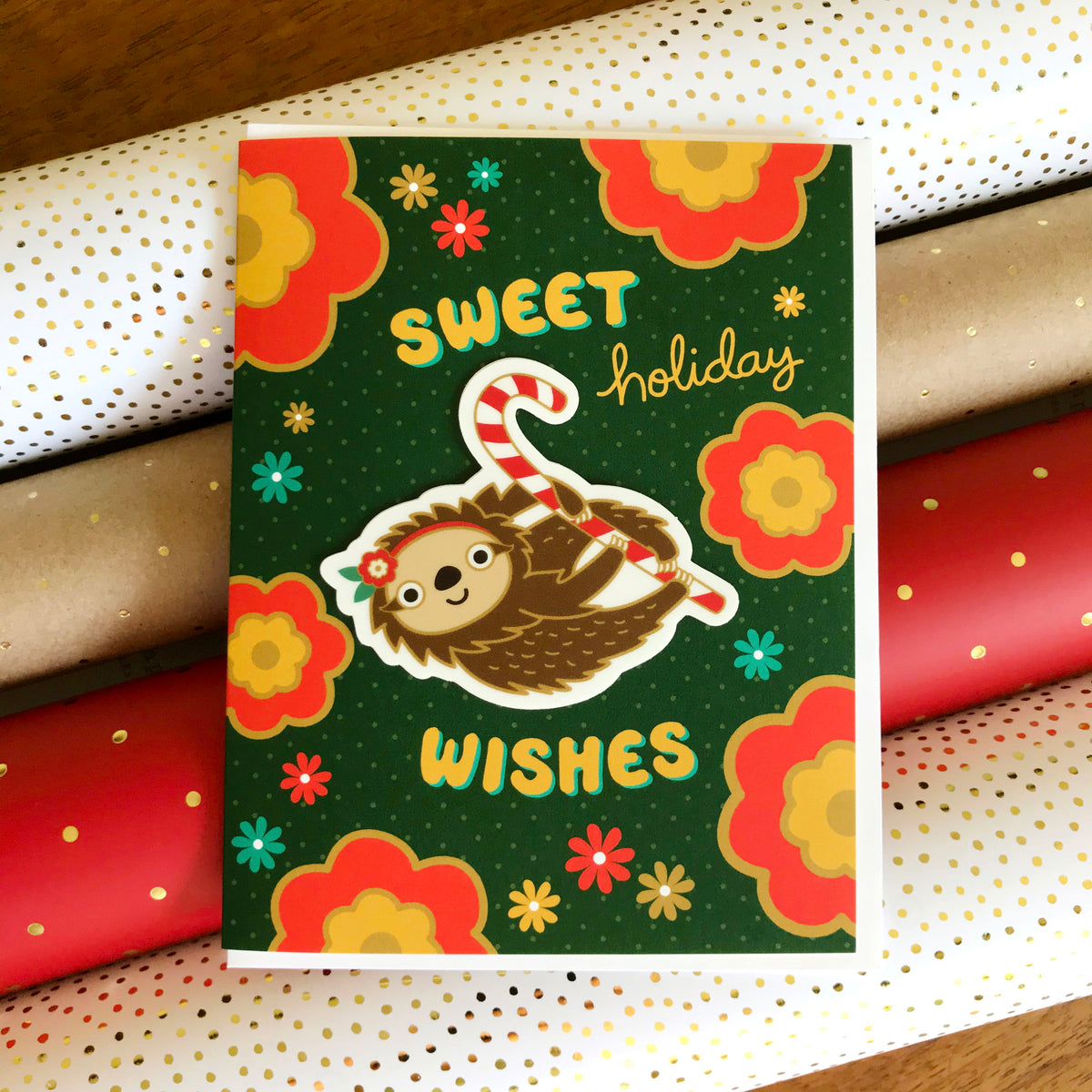 Sweet Sloth Sticker Holiday Card featuring a removable vinyl sloth sticker holding a candy cane on a dark green background with bold red and yellow flowers and the greeting "Sweet Holiday Wishes." Shown with a white envelope. Card sits atop rolls of festive wrapping paper.