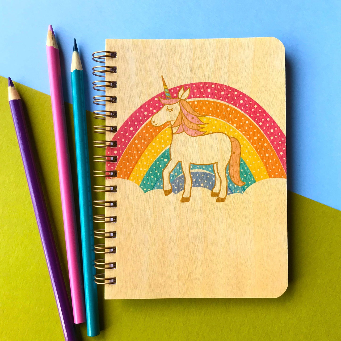 Spiral-bound wood notebook with birch wood covers featuring a unicorn with a pink and orange mane standing in front of a polka-dotted pastel rainbow. Notebook sits on top of colored paper and next to colored pencils.