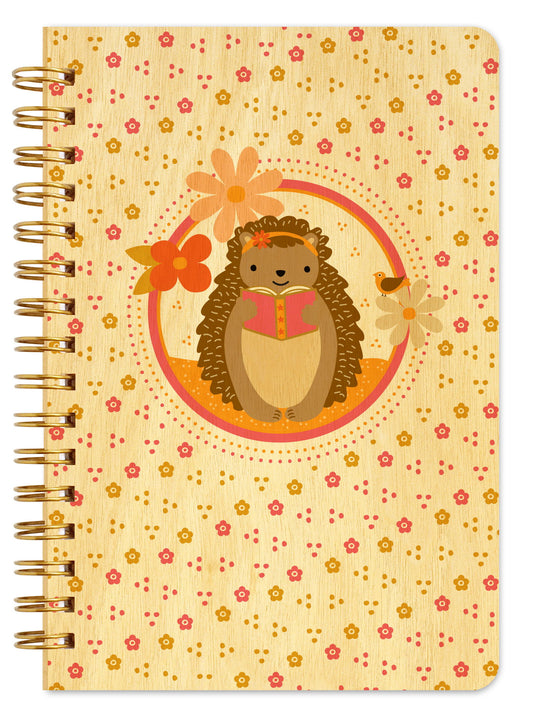 Spiral-bound wood notebook with birch wood covers featuring a reading hedgehog with a floral headband, surrounded by flowers and a small bird inside a circular frame on a floral-patterned background.