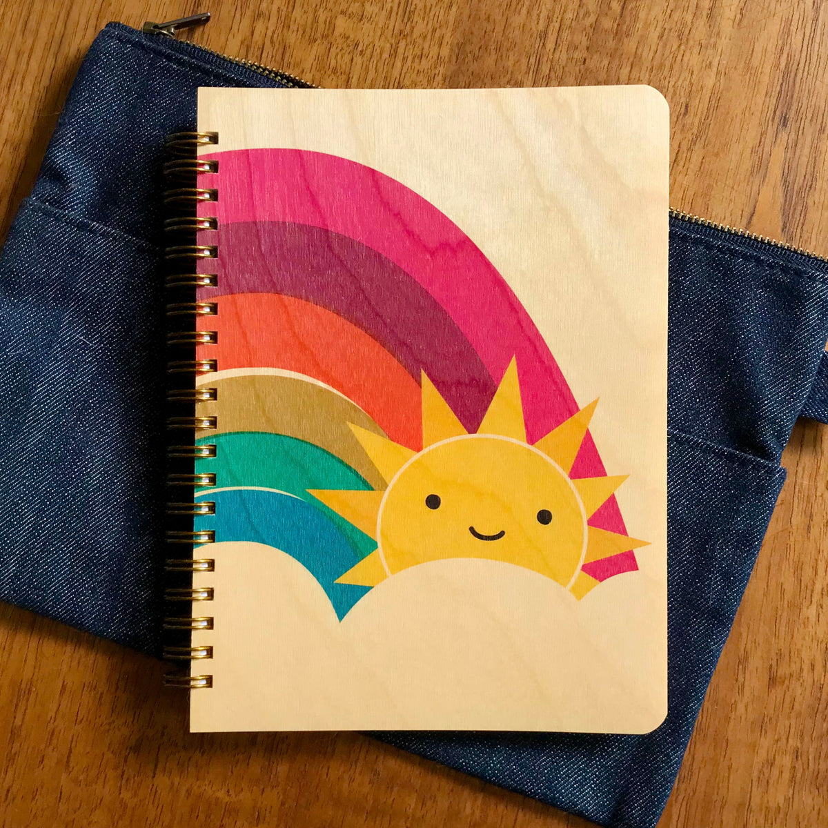Spiral-bound wood journal with birch wood covers featuring a smiling sun peeking out from behind a bold rainbow in pink, orange, gold and teal. Journal sits upon a denim pencil pouch.