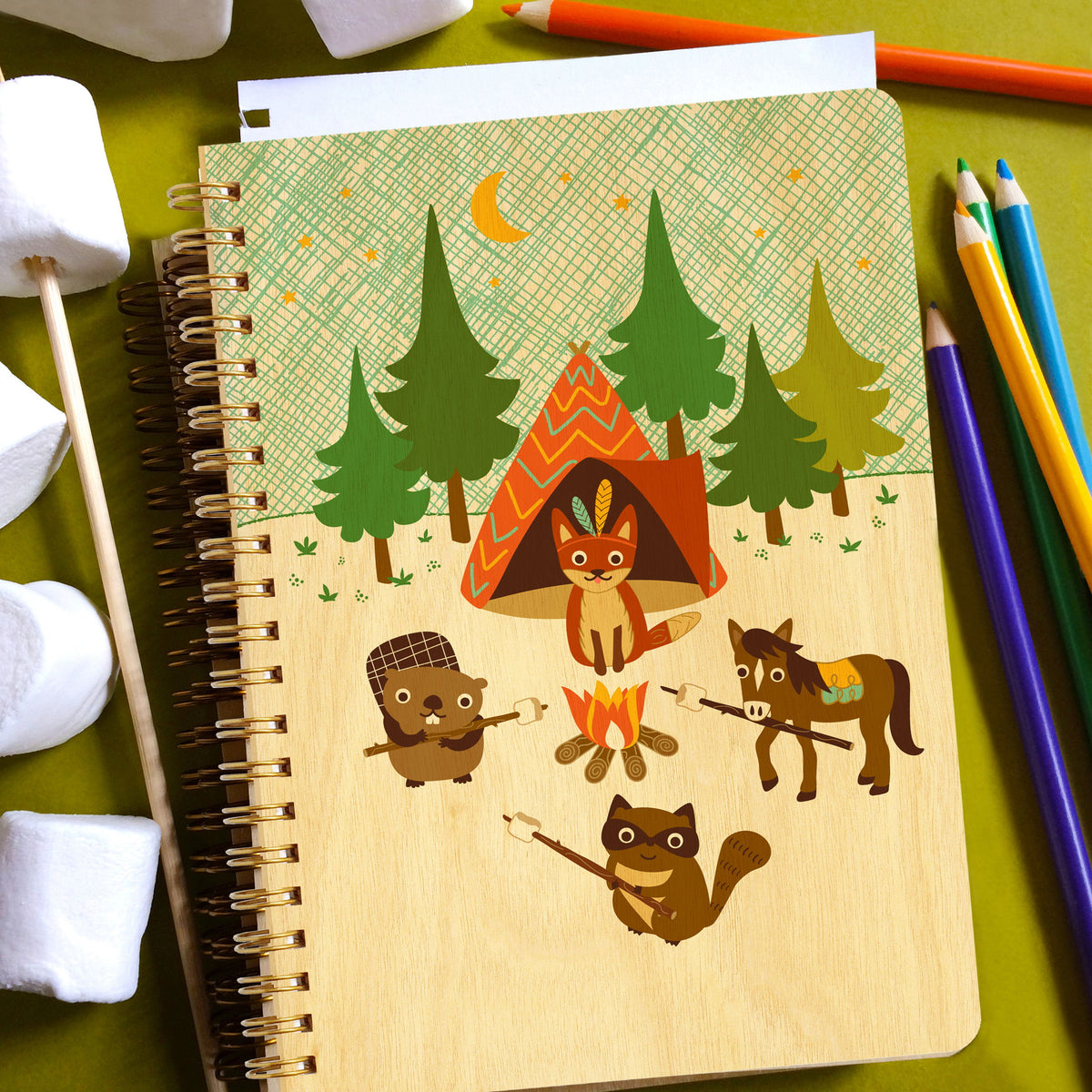 Spiral-bound wood journal with birch wood covers featuring a fox, beaver, horse and raccoon roasting marshmallows around a campfire beside a tent and pine trees under a crescent moon. Journal is accompanied by colored pencils and marshmallows.