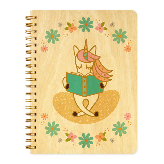 Spiral-bound wood journal with birch wood covers featuring a unicorn with a pink mane absorbed in a teal book, framed by a pastel floral wreath.