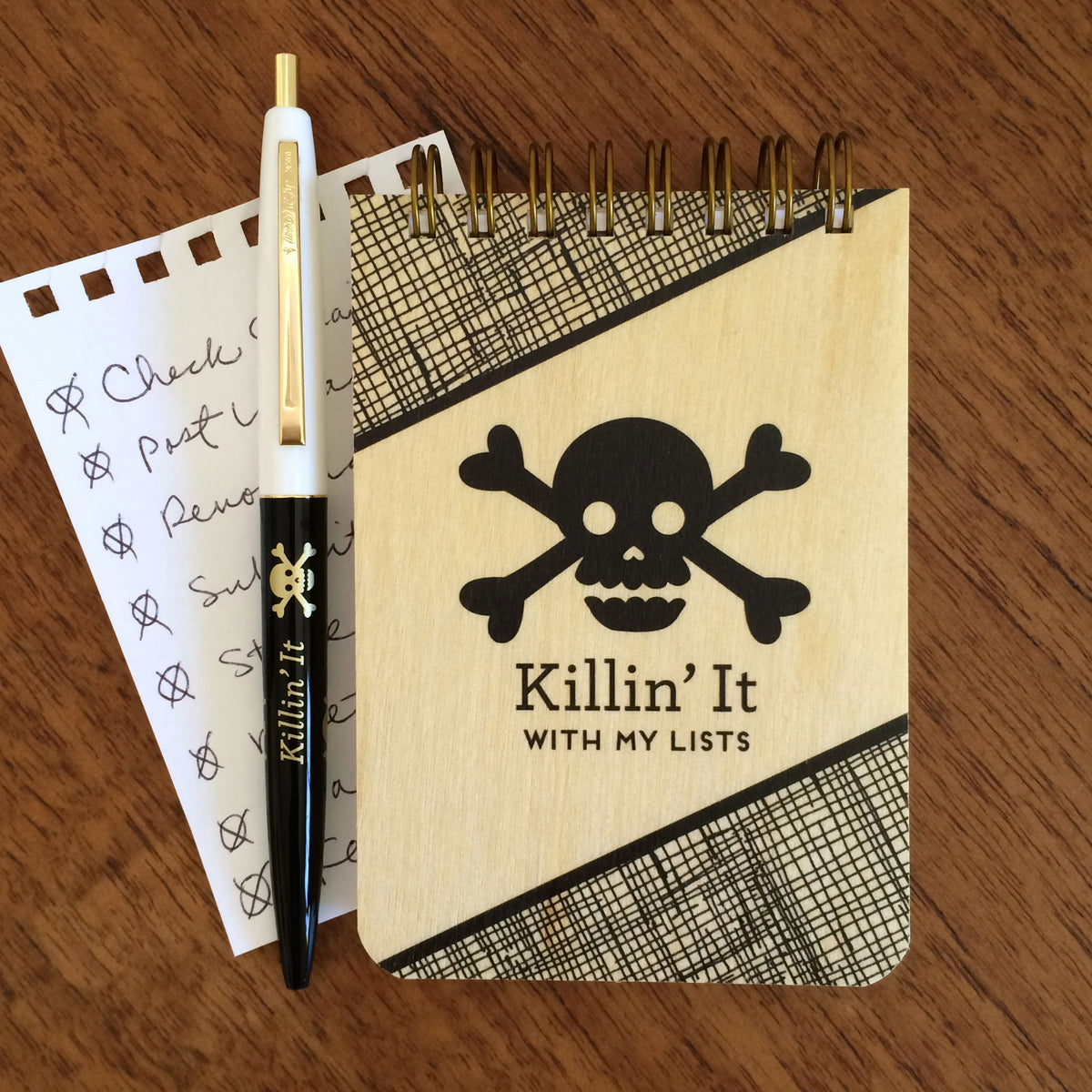 Spiral-bound wood mini notepad with birch wood covers, skull and crossbones design and "Killin' It With My Lists" text. Next to it sits a ballpoint click pen and a torn out sheet of inner paper with a list written on it.
