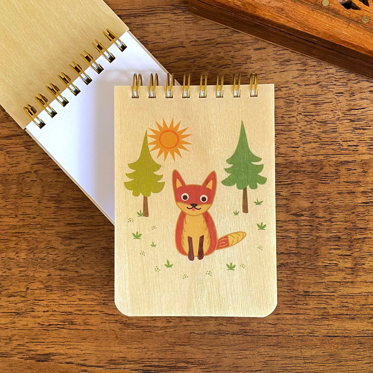Spiral-bound wood mini notepad with birch wood covers featuring a smiling fox sitting in a sunny forest clearing with pine trees sits upon an open mini notepad that shows blank white inner pages.