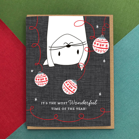 Holiday card pictured on colorful background. Card has a white illustrated cat swinging from the top of the card surrounded by Christmas ornaments on a dark textured background. Text reads 'It's the most wonderful time of the year!'