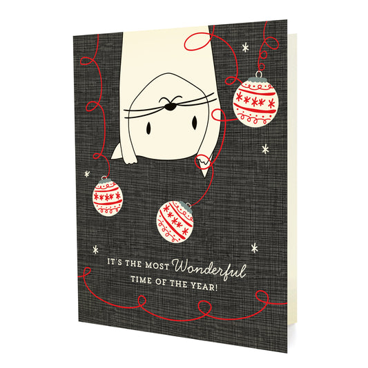 Crazy Kitty folded paper holiday card featuring a white cat dangling upside down and batting at red and cream Christmas ornaments against a dark gray background with the greeting "It's the Most Wonderful Time of the Year!"