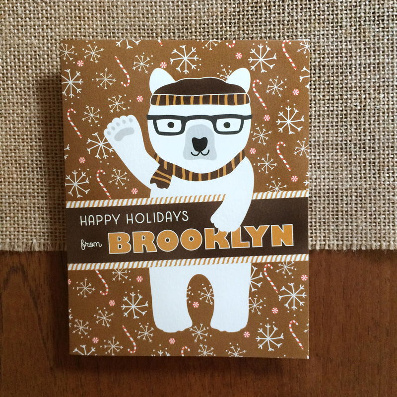 Polar Brooklyn folded paper holiday card featuring a polar bear in black glasses and a brown striped beanie and scarf waving against a warm brown background with snowflakes and candy canes and the greeting "Happy Holidays from Brooklyn"