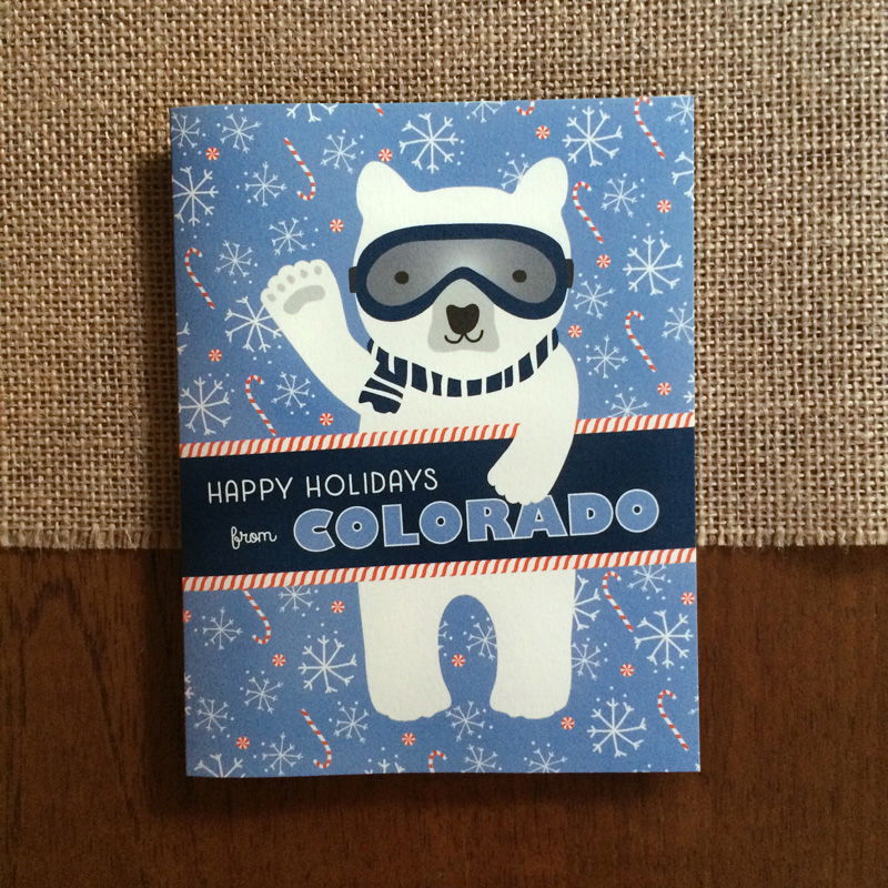 Polar Colorado folded paper holiday card featuring a polar bear in ski goggles and a navy striped scarf waving against a light blue background with snowflakes and candy canes and the greeting "Happy Holidays from Colorado"