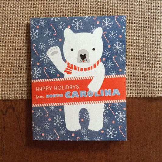 Polar North Carolina folded paper holiday card featuring a polar bear in a red striped scarf waving against a blue-gray background with snowflakes and candy canes and the greeting "Happy Holidays from North Carolina"