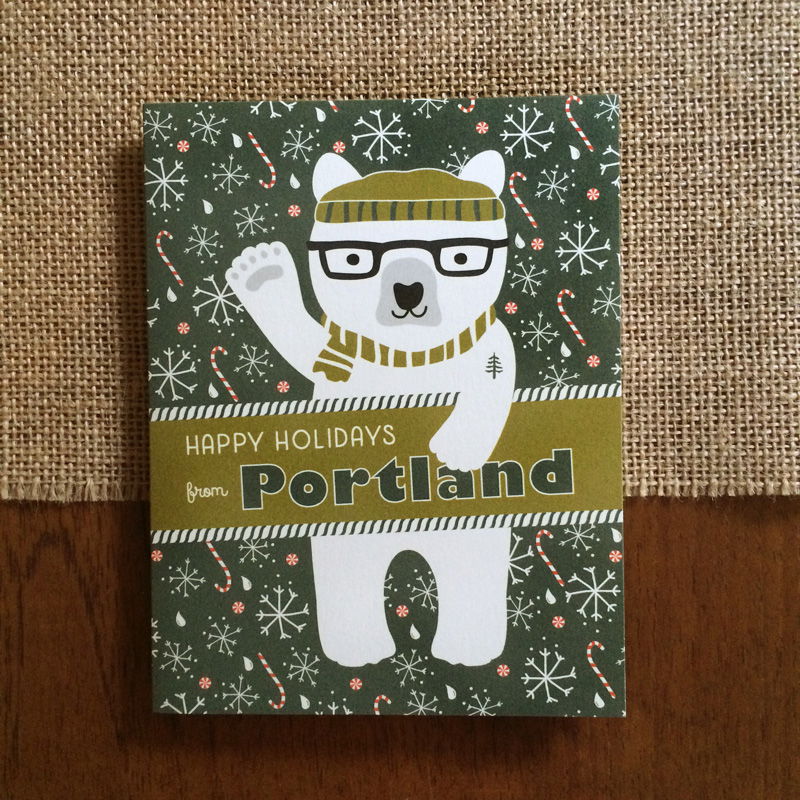 Polar Portland folded paper holiday card featuring a polar bear in black glasses and an olive striped beanie and scarf with a pine tree tattoo waving against a dark green background with snowflakes, raindrops and candy canes and the greeting "Happy Holidays from Portland"