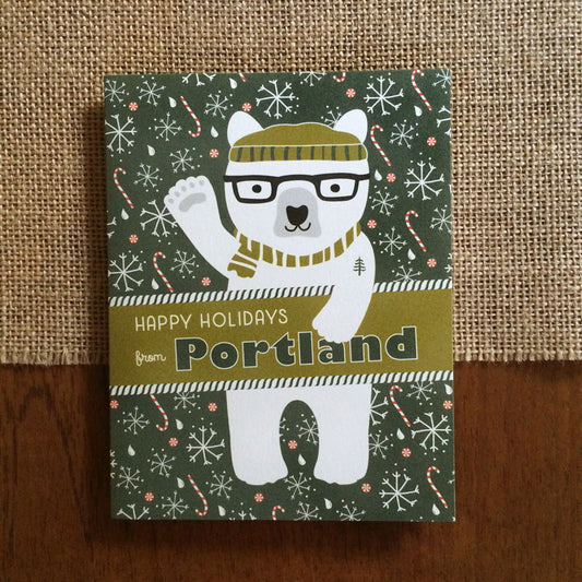 Polar Portland folded paper holiday card featuring a polar bear in black glasses and an olive striped beanie and scarf with a pine tree tattoo waving against a dark green background with snowflakes, raindrops and candy canes and the greeting "Happy Holidays from Portland"