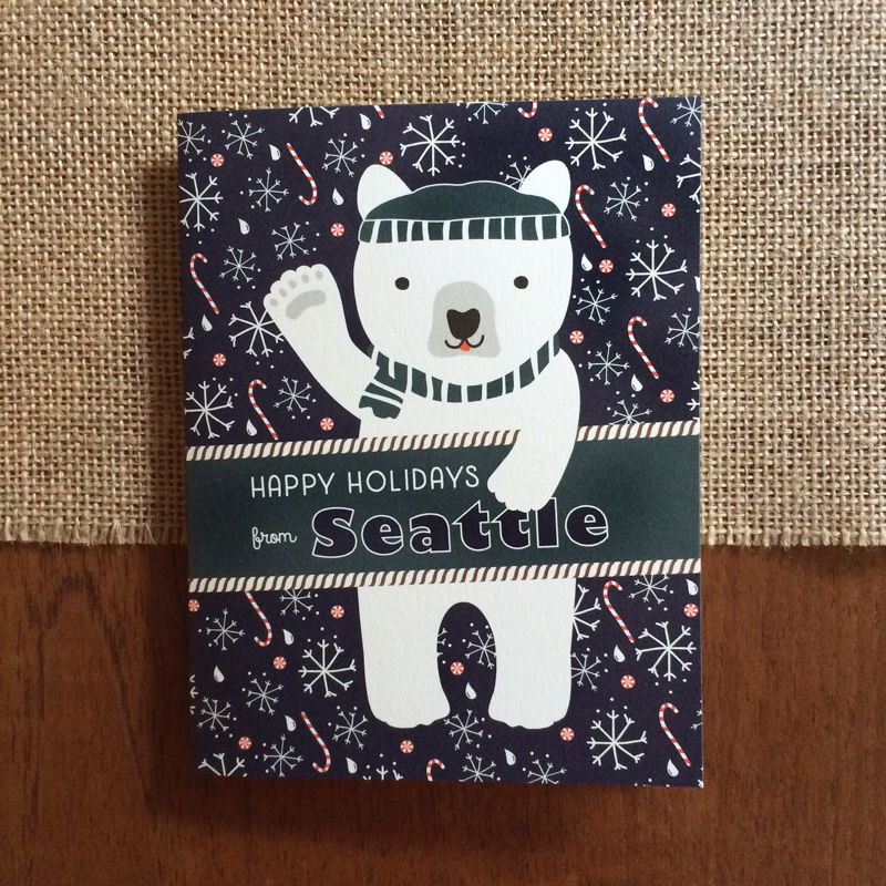 Polar Seattle folded paper holiday card featuring a polar bear in a teal beanie and scarf waving against a navy blue background with snowflakes, raindrops and candy canes and the greeting "Happy Holidays from Seattle"