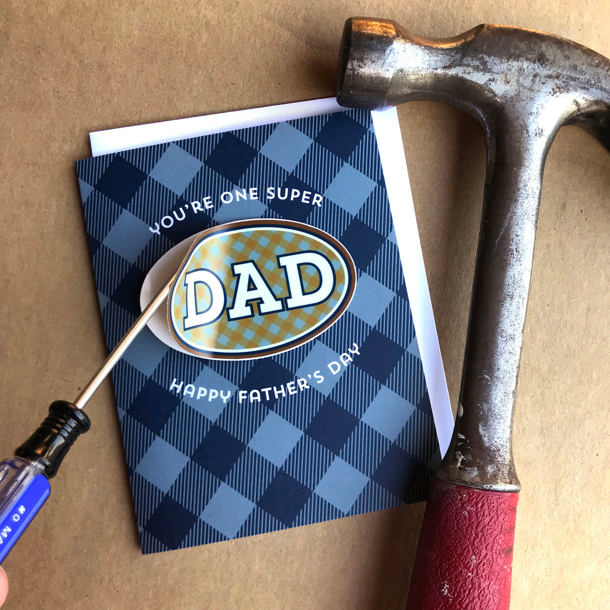 Photo of the Plaid Dad Sticker Father's Day card alongside tools, with a screwdriver lifting up the vinyl sticker, demonstrating that it's easy for recipient to remove.