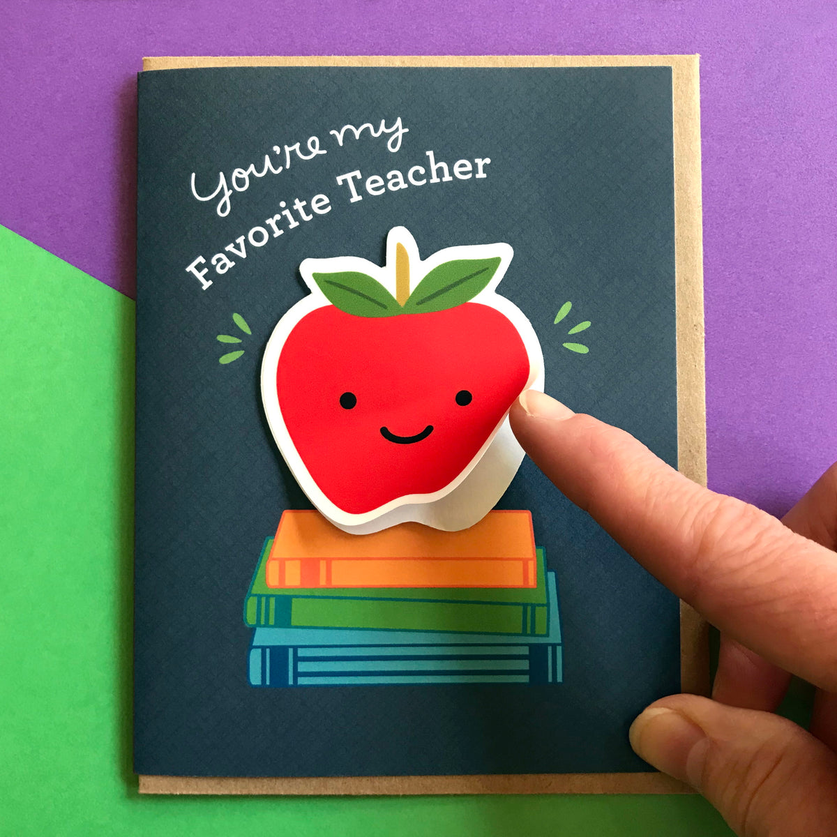 Teacher card featuring an apple on a stack of illustrated books and a finger showing how the vinyl sticker is easy for recipient to remove and use.