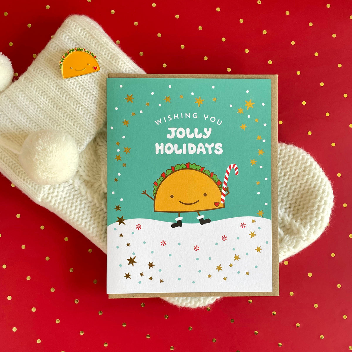 Taco Gold Foil Holiday Card featuring a smiling taco in black boots holding a candy cane against a snowy aqua background with gold foil stars and a "Wishing You Jolly Holidays" greeting. Shown with a kraft envelope next alongside a Taco Lover enamel pin, atop a Christmas stocking on a festive red background.