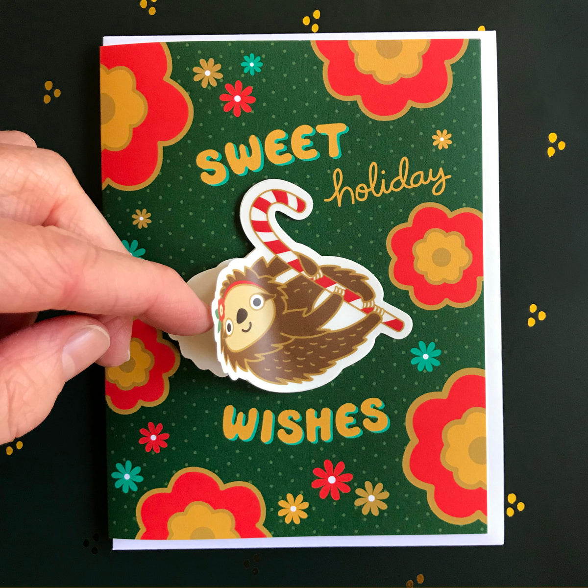 Sweet Sloth Sticker Holiday Card featuring a removable vinyl sloth sticker holding a candy cane on a dark green background with bold red and yellow flowers and the greeting "Sweet Holiday Wishes." Shown with a white envelope. Finger is lifting up the edge of the sticker to show that it's removable.
