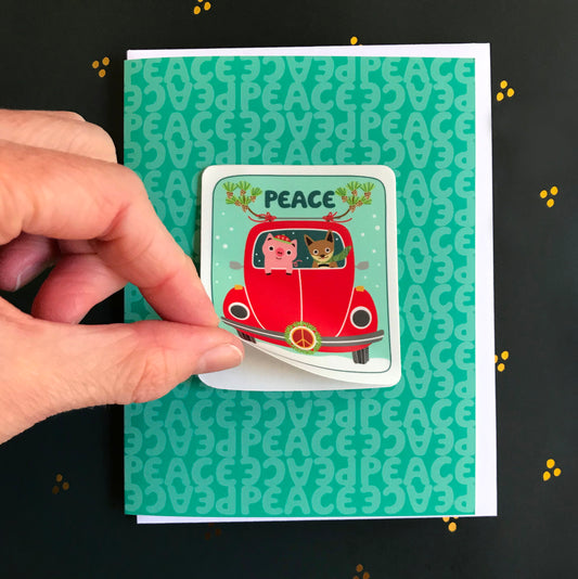Peace Pals Sticker Holiday Card featuring a removable vinyl sticker of a pig and fox driving a red VW bug decorated with pine branches and a peace sign wreath on an aqua background with the word "Peace." Shown with a white envelope. Fingers are shown lifting up the corner of the sticker to demonstrate that it's removable.