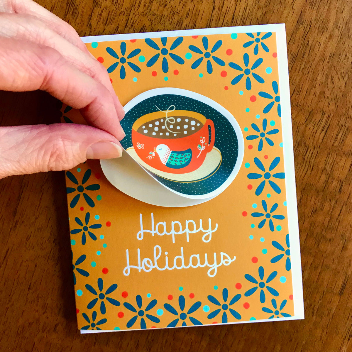 Holiday Cocoa Sticker Holiday Card featuring a removable vinyl sticker of a red mug of hot cocoa with marshmallows and a teal bird design on a warm gold background with teal flowers and the greeting "Happy Holidays." Shown with a white envelope. A finger lifts the edge of the sticker to demonstrate that it's removable.