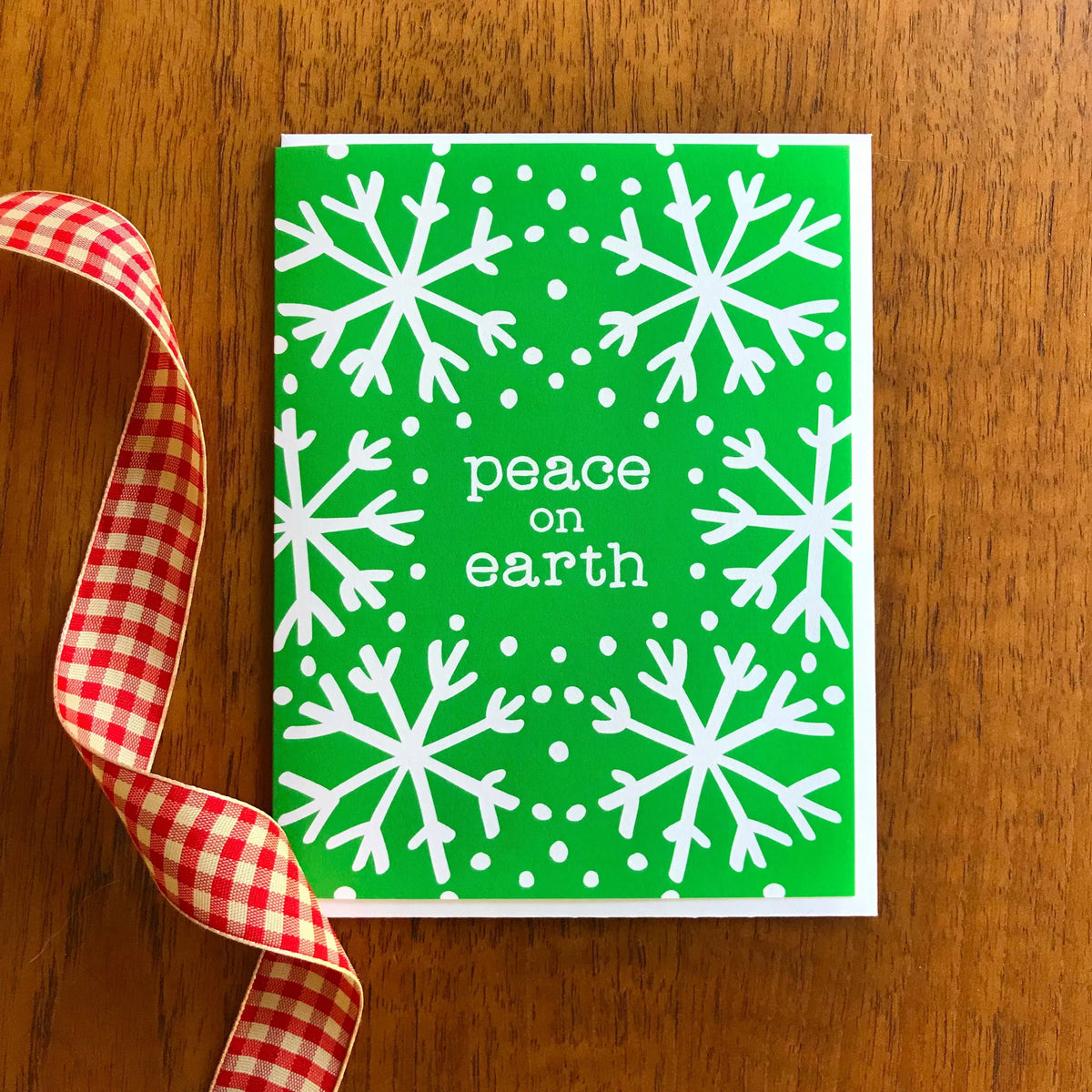 Green Peace folded paper holiday card featuring large white snowflakes on a vivid green background with the greeting "peace on earth." Shown with a white envelope on a wood tabletop.