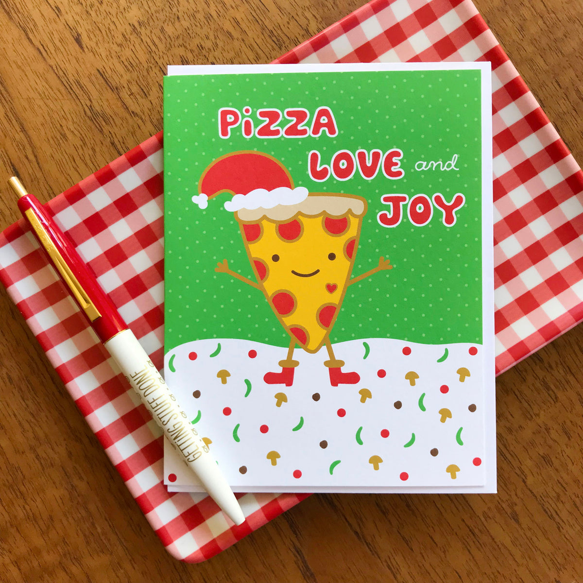 Pizza Love Joy folded paper holiday card featuring a smiling pepperoni pizza slice in a Santa hat and red boots dancing on a snowy ground scattered with mushrooms and peppers against a green background with the greeting "Pizza Love and Joy." Shown with a white envelope and a Getting Stuff Done ballpoint pen, sitting on a red and white checkered plate.