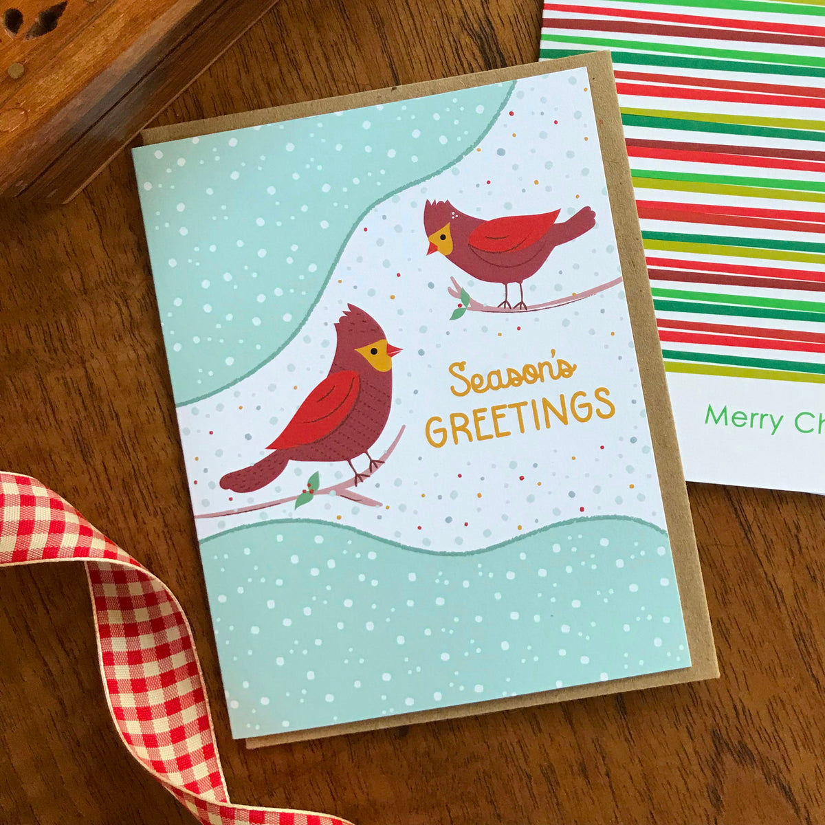 Cardinal Greetings folded paper holiday card featuring two red cardinals perched on branches against a soft mint and white snow-dotted winter background with the greeting "Season's Greetings." Shown with a kraft envelope next to the Simply Merry holiday card on a wood tabletop.
