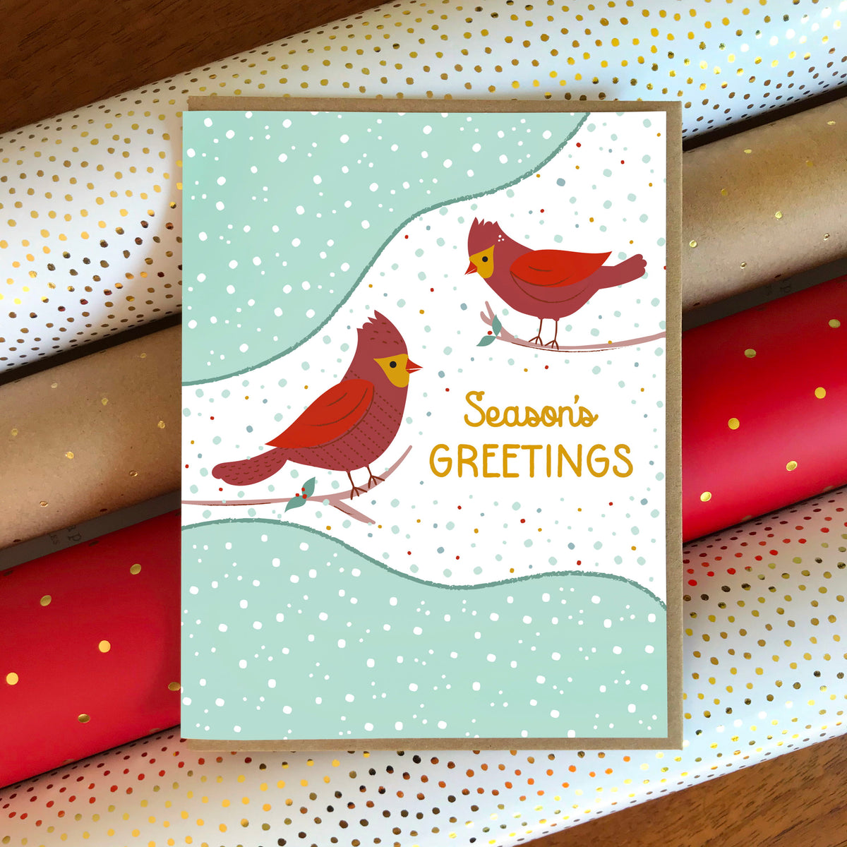 Cardinal Greetings folded paper holiday card featuring two red cardinals perched on branches against a soft mint and white snow-dotted winter background with the greeting "Season's Greetings." Shown with a kraft envelope atop rolls of festive wrapping paper.