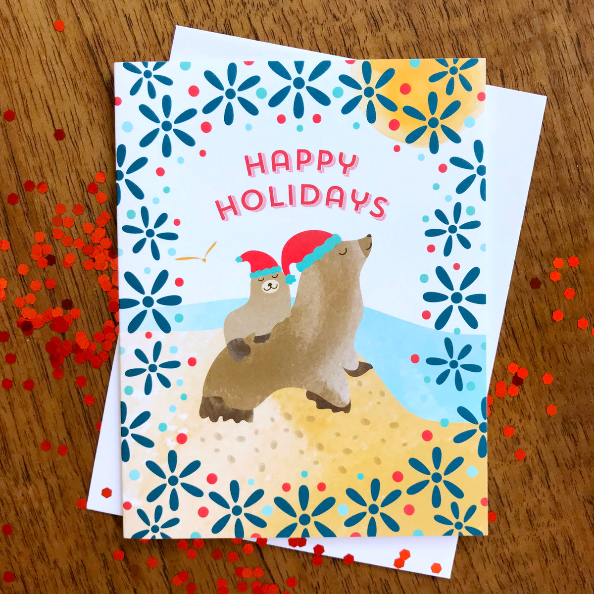 Sea Lions folded paper holiday card featuring two sea lions in red Santa hats on a sandy beach with aqua ocean waves, navy blue snowflake florals and a "Happy Holidays" greeting. Shown with a white envelope on a wood tabletop.