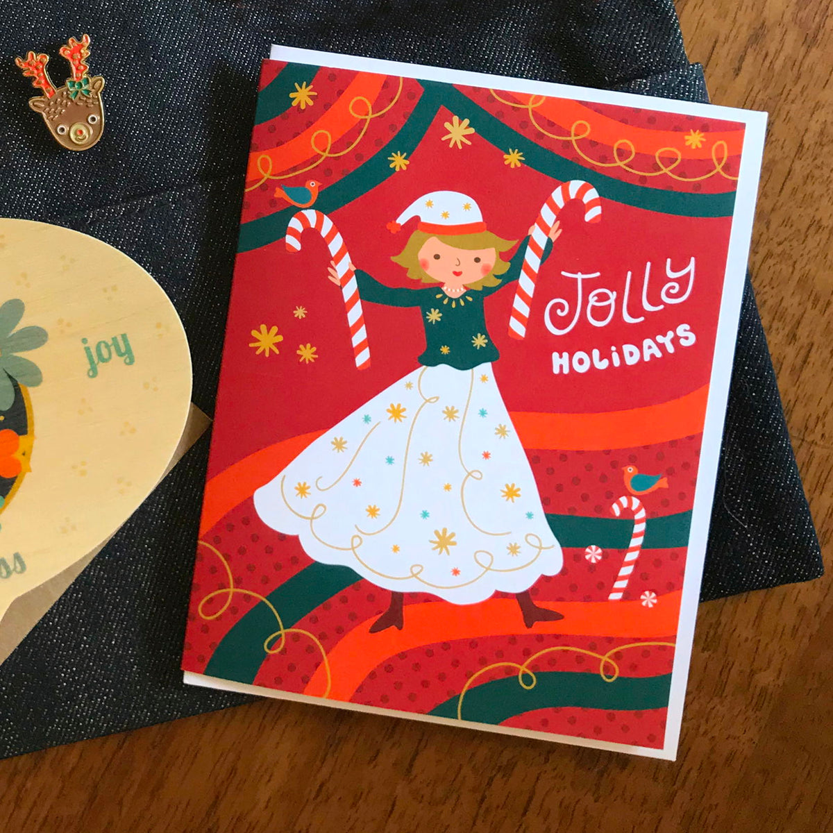 Jolly Gal folded paper holiday card featuring a dancing woman in a white dress and Santa hat holding candy canes against a bold red and green swirling background with colorful birds and the greeting "Jolly Holidays." Shown with a white envelope. A reindeer enamel pin sits nearby.